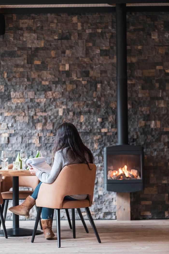 A woman sits in a restaurant reading the menu in a cozy atmosphere, with a fire burning in the fireplace in the background.