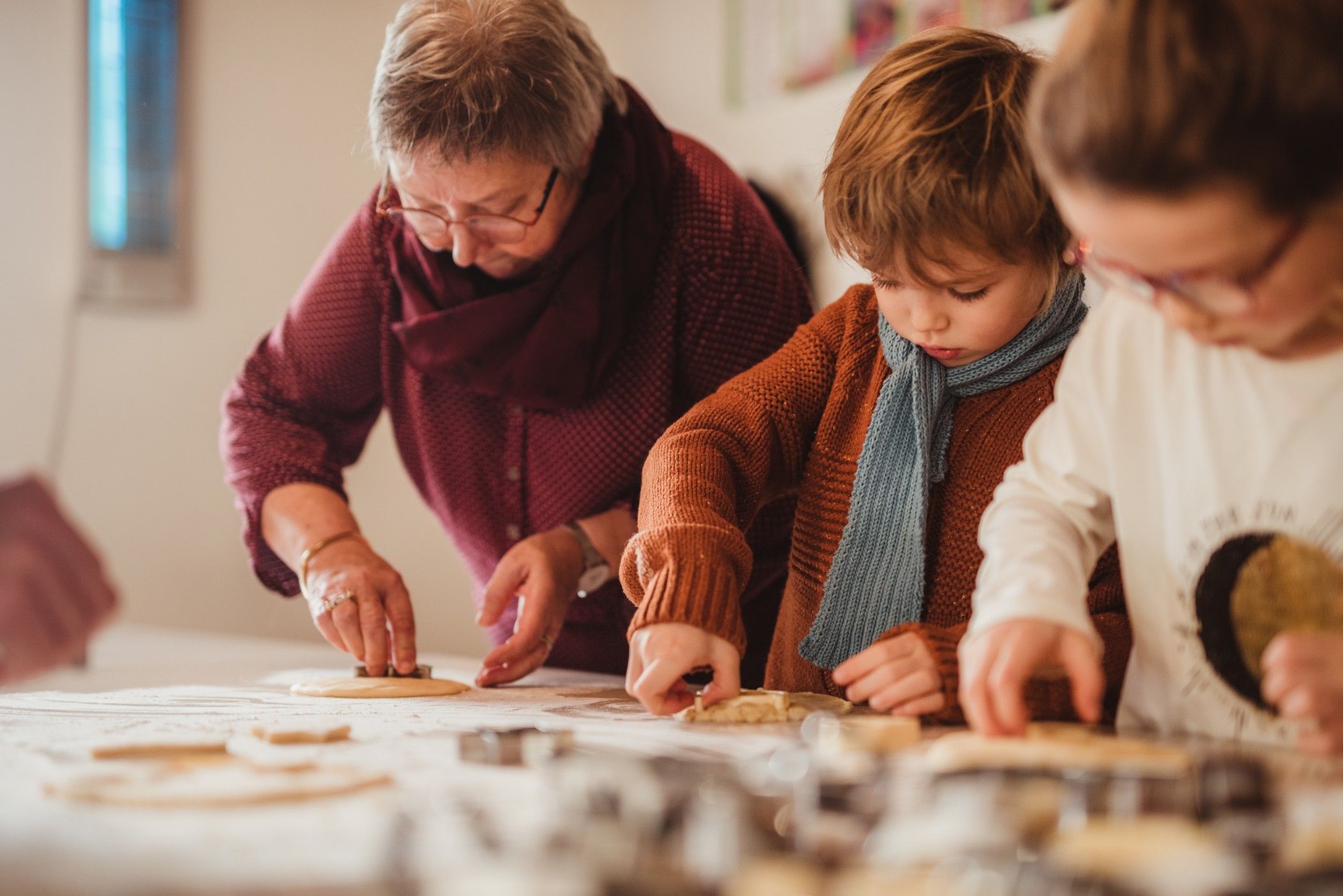 Children and a woman sit at a table, baking cookies together, while the children use cookie cutters to cut out shapes.