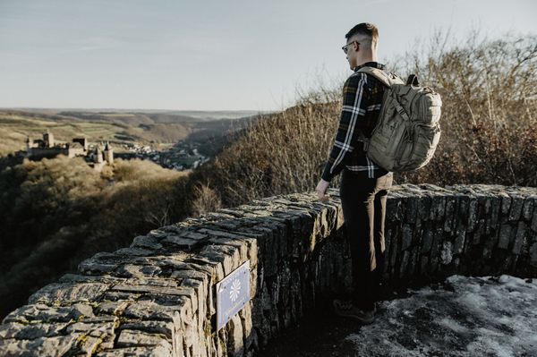 Ein Mann steht mit Rucksack auf einer Aussichtsplattform aus Stein und schaut auf ein Schloss.