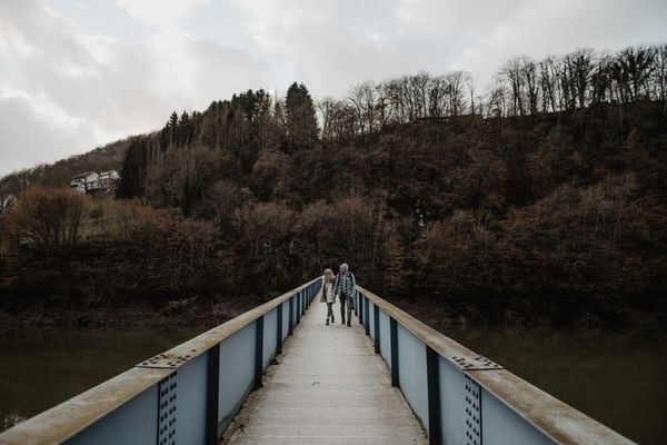 A couple strolls across a pedestrian bridge with a forest in the background.