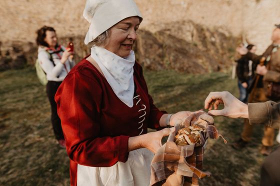 Een vrouw in een rood jurk en wit schort serveert eten aan een groep bezoekers voor een kasteel.